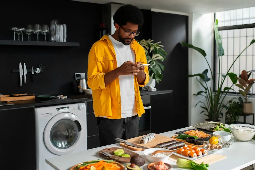 Man preparing sushi in a modern, well-lit kitchen with black walls and indoor plants, using fresh ingredients and a bamboo rolling mat, while referencing his phone for instructions
